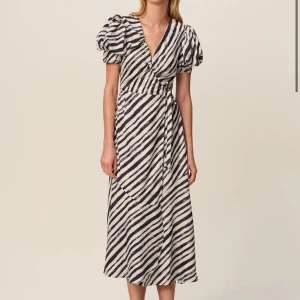 Randig omlottklänning med puffärm - Snygg omlottklänning med svartvita diagonala ränder och puffiga korta ärmar. Klänningen har v-ringning, knyt i midjan och en luftig, lång siluett. Perfekt för dig som gillar en trendig och bekväm stil.