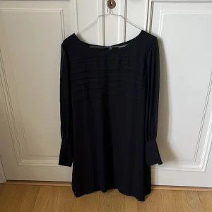 Svart långärmad klänning  - Stilren svart  med lång ärm och diskreta veckdetaljer över bröstet. Klänningen har en rak, något lös passform och är tillverkad i ett mjukt, lätt material som faller snyggt. Perfekt för dig som gillar minimalistisk och tidlös stil.