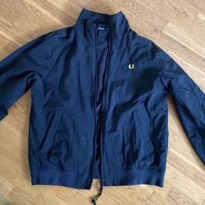Fredperry vindjacka M - Snygg svart bomberjacka från Fred Perry med klassisk gul logga på bröstet. Jackan har ståkrage, dragkedja framtill och två fickor. Materialet är slätt och lätt, perfekt för dig som vill ha en stilren och tidlös look.