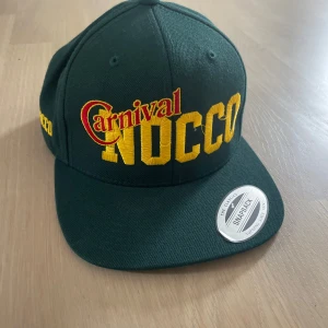  NOCCO Carnival keps  - Mörkgrön snapback keps från NOCCO med broderad Carnival-logga i gult och rött framtill. Klassisk flat brim och justerbar baksida. Snygg streetstyle med tydlig branding och cool färgkombination. Perfekt accessoar för dig som gillar sportiga vibes. Har bara andvänts typ 2 gånger och känns som helt ny