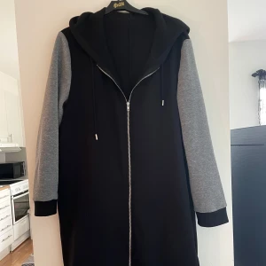 Svart lång hoodiejacka med grå ärmar - Lång hoodiejacka i svart med kontrasterande grå ärmar och svarta muddar. Jackan har huva med dragsko och hel dragkedja framtill. Perfekt för chill dagar och streetstyle. Mjuk och skön bomullskänsla.