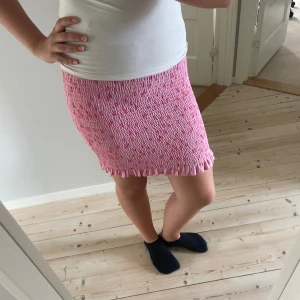 Rosa blommig kortkjol från Pieces - Supersöt rosa kortkjol från Pieces med små blommor i rött och gult. Kjolen är smockad och har volangkant nertill för en gullig vibe. Perfekt för dig som gillar färg och mönster! Materialet är mjukt och stretchigt.