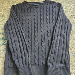 Mörkgrå kabelstickad tröja GANT - Säljer en mörkblå kabelstickad tröja från GANT i storlek S. Tröjan har rund halsringning, ribbade muddar och GANT logga på bröstet. Perfekt för dig som gillar klassisk och stilren look med lite preppy känsla.