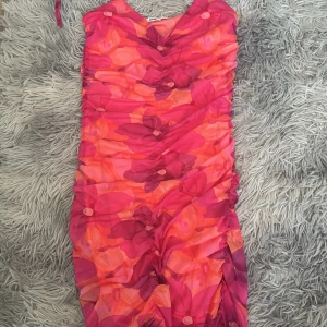 Blommig kortklänning från Sinsay XS - Supersöt kortklänning från Sinsay i storlek XS med blommigt mönster i rosa, rött och orange toner. Klänningen har smala axelband, v-ringning och rynkade detaljer framtill. Perfekt för dig som vill sticka ut med färg och mönster!