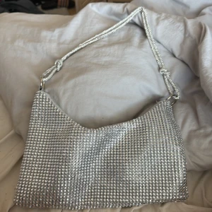 Silvrig axelväska med paljetter - Snygg silvrig axelväska täckt med glittrande paljetter. Perfekt för att ge en extra touch till din outfit. Väskan har en enkel design med en justerbar axelrem och ett rymligt inre fack.