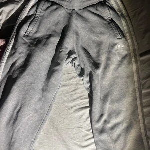 Grå mjukisbyxor från Adidas - Säljer ett par grå mjukisbyxor från Adidas med klassiska ränder längs sidorna. Byxorna har en bekväm passform med resår i midjan och muddar vid bensluten. Perfekta för en avslappnad stil.