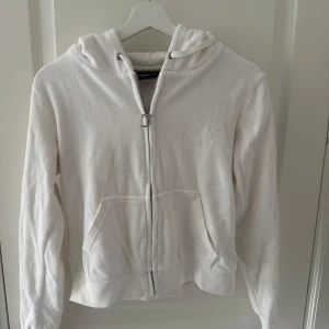 Vit hoodie från Juicy Couture - Säljer en stilren vit hoodie från Juicy Couture med dragkedja och broderad logga på bröstet. Tröjan har en klassisk design med huva och fickor framtill. Perfekt för en avslappnad look.