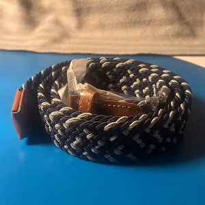 Ny Flätat bälte  - Snyggt flätat bälte i blått och vitt med bruna läderdetaljer. Perfekt för att ge en stilren touch till din outfit. Bältet har ett klassiskt spänne och är tillverkat i ett slitstarkt material. Bältet har aldrig blivit använd den kommer med og förpackning 😊