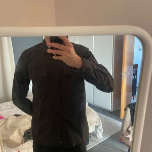 Svart overshirt med knappar - Snygg svart overshirt med knappar framtill. Perfekt för lager-på-lager-stil. Långärmad design med klassisk krage och bröstfickor.