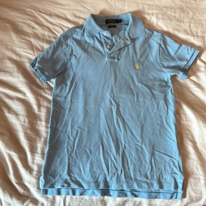 Ljusblå pikétröja från Ralph Lauren - Säljer en stilren ljusblå pikétröja från Ralph Lauren. Tröjan har en klassisk krage och korta ärmar med en broderad gul logotyp på bröstet. Perfekt för en avslappnad men ändå snygg look. Slim fit passform för en modern siluett.