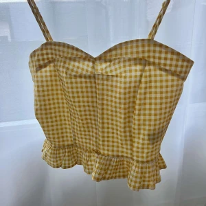 Gul och vit rutig linnetopp - Säljer en söt gul och vit rutig linnetopp med smala axelband och volangdetalj vid nederkanten. Toppen har en dragkedja i ryggen för enkel på- och avtagning. Perfekt för sommarens varma dagar! 🌼