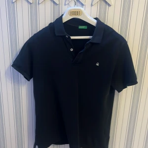 Svart pikétröja från United Colors of Benetton - Modellen är 173 och 60 kg. Snygg svart pikétröja från United Colors of Benetton med korta ärmar och klassisk krage. Tröjan har en liten broderad logga på bröstet och knappar vid halsen. Perfekt för en stilren look.