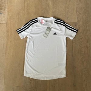 Vit t-shirt från Adidas - Säljer en vit t-shirt från Adidas med klassiska svarta ränder längs ärmarna och Adidas-logga på bröstet. Perfekt för träning eller en sportig look. Kortärmad och tillverkad i ett lätt och bekvämt material.
