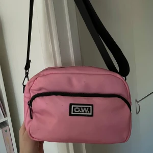 Rosa axelväska från O.W. - Säljer en söt rosa axelväska från O.W. med svart dragkedja och justerbar axelrem. Perfekt för att bära dina nödvändigheter med stil. Väskan har en praktisk storlek och ett enkelt men trendigt utseende.