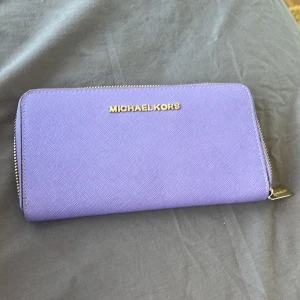 Lila plånbok från Michael Kors - Snygg lila plånbok från Michael Kors med dragkedja runtom. Tillverkad i ett slitstarkt syntetmaterial och har flera fack för kort och kontanter. Perfekt för att hålla ordning på dina värdesaker med stil.