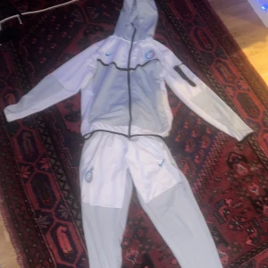 Vit och grå träningsjacka från Nike - Säljer en stilren vit och grå träningsjacka från Nike med dragkedja och huva. Jackan har långa ärmar och en praktisk ficka på ärmen. Perfekt för sportiga aktiviteter och träning.