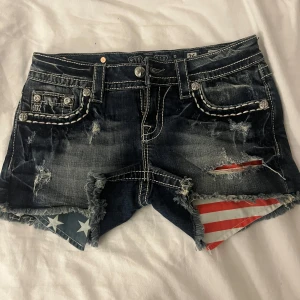 Jeansshorts från Miss Me - Snygga jeansshorts från Miss Me med slitna detaljer och fransar. De har en cool amerikansk flagga som sticker fram vid fickorna och dekorativa sömmar på bakfickorna. Perfekta för en avslappnad stil. Helt nya, oanvända, endast testade. 