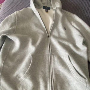 Grå hoodie från Ralph Lauren - Säljer en klassisk grå hoodie från Ralph Lauren med dragkedja och fickor framtill. Perfekt för en avslappnad stil. Den har en liten logga på bröstet och är tillverkad i ett mjukt material.