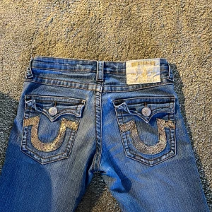Blå jeans med detaljer från True Religion - Snygga blå jeans från True Religion med unika detaljer på bakfickorna. Byxorna har en klassisk femficksdesign och knappar med logotyp. Perfekta för dig som vill ha något extra i garderoben.