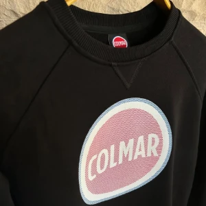 Svart sweatshirt från Colmar - Snygg svart sweatshirt från Colmar med ett stort, runt logotryck i vitt och rosa på framsidan. Tröjan har långa ärmar och ribbade muddar vid ärmslut och nederkant. Perfekt för en avslappnad stil.
