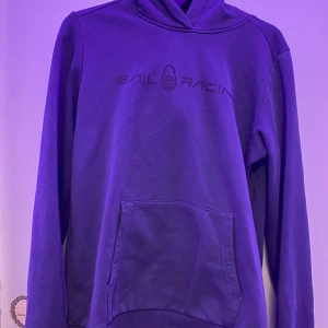Blå hoodie från Sail Racing - Säljer en snygg blå hoodie från Sail Racing. Den har en klassisk design med en stor ficka framtill och en bekväm huva. Perfekt för kyliga dagar och avslappnade stunder. Passar både för vardag och fritid.