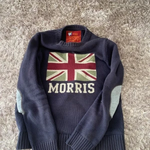 Mörkblå stickad tröja från Morris - Säljer en snygg mörkblå stickad tröja från Morris med Union Jack-motiv och texten 'Morris' på framsidan. Tröjan har långa ärmar och grå armbågslappar som ger en stilren touch. Perfekt för en avslappnad och trendig look. Väldigt bra skick