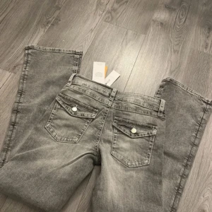 Grå jeans med vida ben - Säljer ett par grå jeans från Name It med vida ben och klassisk femficksdesign. Jeansen har en knapp och dragkedja framtill samt dekorativa knappar på bakfickorna. Perfekta för en avslappnad stil.