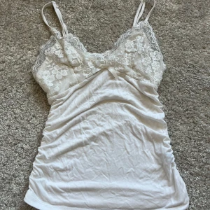 Vit spetslinne med blommönster - Säljer ett vackert vitt linne med spetsdetaljer och blommönster. Linnet har smala axelband och en söt rosett framtill. Perfekt för en romantisk look eller som ett lager under andra plagg. Använd fåtal gånger 💕