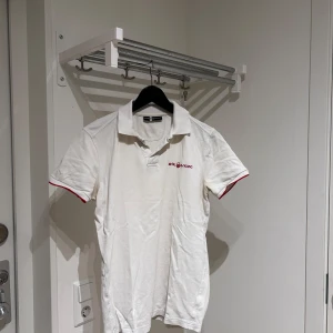 Vit kortärmad pikétröja från sail racing - Säljer en stilren vit pikétröja från Tommy Hilfiger med röda detaljer på ärmarna och logotypen broderad på bröstet. Perfekt för en avslappnad men ändå snygg look. Passar bra till både jeans och shorts.