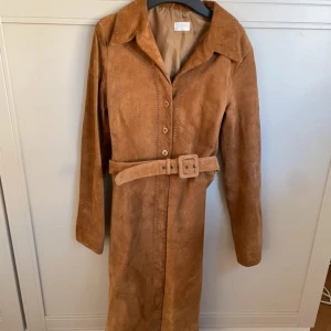 Brun mockakappa med bälte - Säljer en elegant brun mockakappa med knappar och ett matchande bälte i midjan. Kappan har långa ärmar och en klassisk krage. Perfekt för höst och vår!