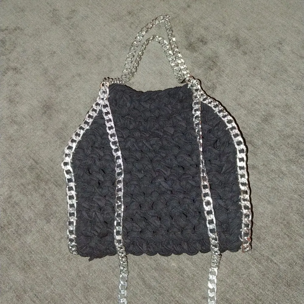 Snygg svart virkad väska med silverfärgade kedjedetaljer. Perfekt för att ge din outfit en unik touch. Väskan har en enkel design med två kedjehandtag som ger en cool kontrast till det mjuka materialet.. Laukut & Käsilaukut.