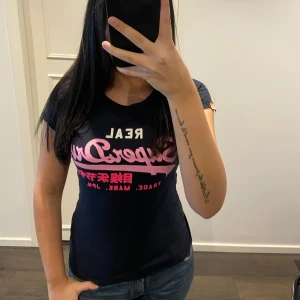 Svart t-shirt från Superdry - Snygg svart t-shirt från Superdry med rosa och vit logga på bröstet. Perfekt för en casual look. T-shirten har en perfekt passform och korta ärmar.