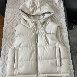 Beige/vit dunväst med huva - Snygg beige dunväst med dragkedja och huva. Perfekt för kyligare dagar. Västen har en stilren design med praktiska fickor och dragsko i huvan.