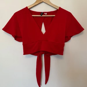 Röd omlottopp från H&M - Snygg röd omlottopp från H&M med korta ärmar och knytning i midjan. Toppen har en v-ringad design och öppen rygg.