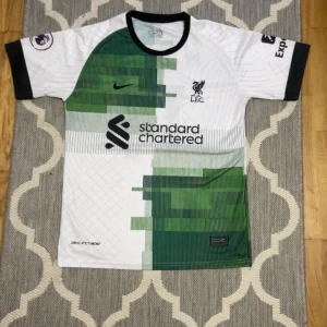 Vit och grön fotbollströja från Nike l.f.c MOHAMMED SALAH - Säljer en cool vit och grön fotbollströja från Nike med svart krage och ärmslut. Tröjan har ett unikt mönster och klubbens logga på bröstet. Perfekt för fotbollsfans! 💚⚽