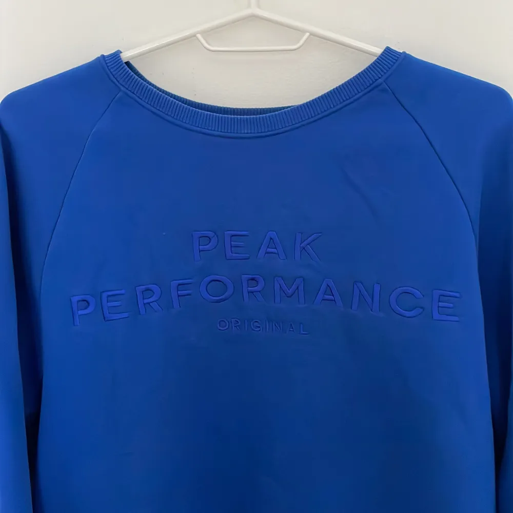 Säljer en snygg blå tröja från Peak Performance med broderad logga på bröstet. Tröjan har långa ärmar och en rund halsringning. Perfekt för en stilren och sportig look.. Neuletakit & Villapaidat.