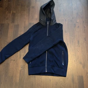 Mörkblå jacka från Ralph Laurent (RLX) - Snygg mörkblå jacka från Ralph Lauren (RLX) med huva och dragkedja. Jackan har ribbade detaljer på ärmarna och en stilren design. Perfekt för kyligare dagar. Den är i storlek M. Nypris runt 6000 kr.