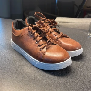 Bruna sneakers med vit sula - Snygga bruna sneakers i skinn med vit sula och snörning. Perfekta för en stilren look. De har en rund tå och en klassisk design som passar till många olika outfits.