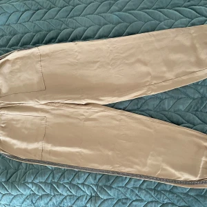 Beige byxor från Zara - Snygga beige byxor från Zara med elastisk midja och glittriga detaljer längs sidorna. Perfekta för en stilren look med en touch av glamour.