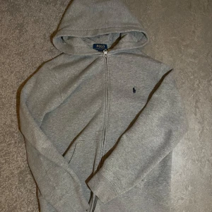 Grå hoodie från Ralph Lauren - Säljer en stilren grå hoodie från Ralph Lauren med dragkedja och huva. Tröjan har en klassisk design med det ikoniska logotypbroderiet på bröstet. Perfekt för en avslappnad look. Storlek L 150-161 cm i junior 
