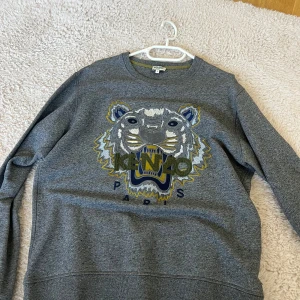 Grå sweatshirt från Kenzo med tigerbroderi - Säljer en grå sweatshirt från Kenzo med ett stort, broderat tigerhuvud i mitten. Tröjan har rund hals och långa ärmar. Perfekt för en avslappnad och stilren look.