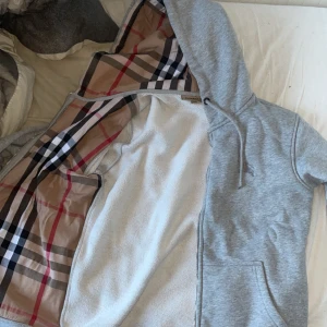 Grå hoodie med mönstrat foder - Säljer en stilren grå hoodie med ett klassiskt rutigt mönster på insidan. Den har en broderad logga på bröstet och snörning vid huvan. Perfekt för en avslappnad stil. Ena snöret har tappat metal biten som sitter på den men syns knappt dragkedjan är sönder!
