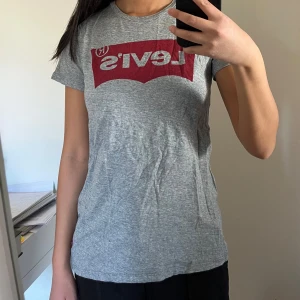 Grå t-shirt från Levi's - Säljer en klassisk grå t-shirt från Levi's med röd logga på bröstet. T-shirten har en normal passform och korta ärmar, perfekt för en avslappnad stil. Passar till både jeans och shorts! Funkar även för nån som är storlek S enligt mig. 