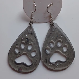 Grå örhängen med tassmotiv - Dessa är 2a utrensning! Unika grå örhängen i form av droppar med utskuret tassmotiv. De har en glittrig yta med små inslag av silver och svart. Perfekta för djurälskare som vill ha något speciellt i sin smyckessamling.