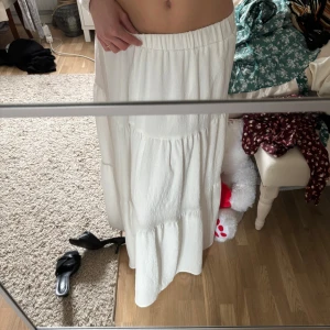 Vit långkjol - Säljer en stilren vit långkjol med elastisk midja. Kjolen har en luftig passform och är perfekt för varma dagar. Den är tillverkad i ett lätt och bekvämt material som ger en avslappnad look. Skulle rekommendera kjolen till folk kortare än 165cm, då går den ner till fötterna💕