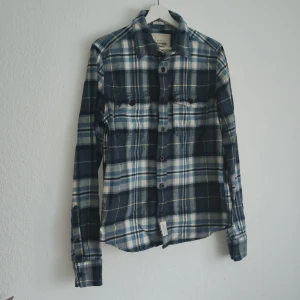 Rutig skjorta från Abercrombie & Fitch - Säljer en stilren rutig skjorta från Abercrombie & Fitch i blått, vitt och gult. Skjortan har långa ärmar och knappar framtill. Perfekt för en avslappnad look.