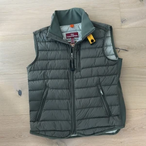 Grön dunväst från Parajumpers - Snygg mörkgrön dunväst från Parajumpers med quiltad design och hög krage. Västen har dragkedja framtill, tre fickor med dragkedja och den klassiska gula Parajumpers-detaljen vid halsen. Perfekt lager-på-lager-plagg för kyliga dagar.