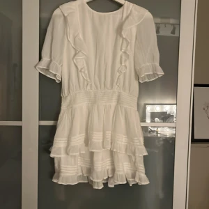 Vit volangklänning från Gina Tricot - Supersöt vit kortklänning från Gina Tricot med volanger, korta ärmar och knytning i nacken. Klänningen har flera lager volanger på kjolen och fina detaljer längs bröstet. Perfekt för sommarens alla tillfällen!