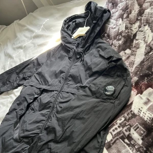  CP Company jacka  - Äkta Svart vindjacka från C.P. Company med huva och dragkedja framtill. Jackan har den ikoniska linsen på ärmen och justerbar dragsko vid huvan. Tillverkad i lätt polyester, perfekt för blåsiga dagar. Snygg och stilren design som passar casual, 