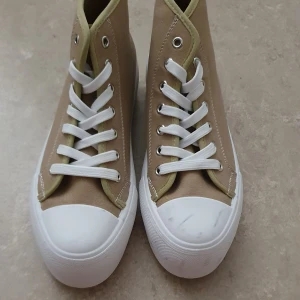 Sneakers - Stilrena höga sneakers i beige canvas med vita gummisulor och tåhätta. Skorna har vita snören och silverfärgade öljetter, samt en svart rand längs sulan. Perfekta för dig som gillar en clean och klassisk look.